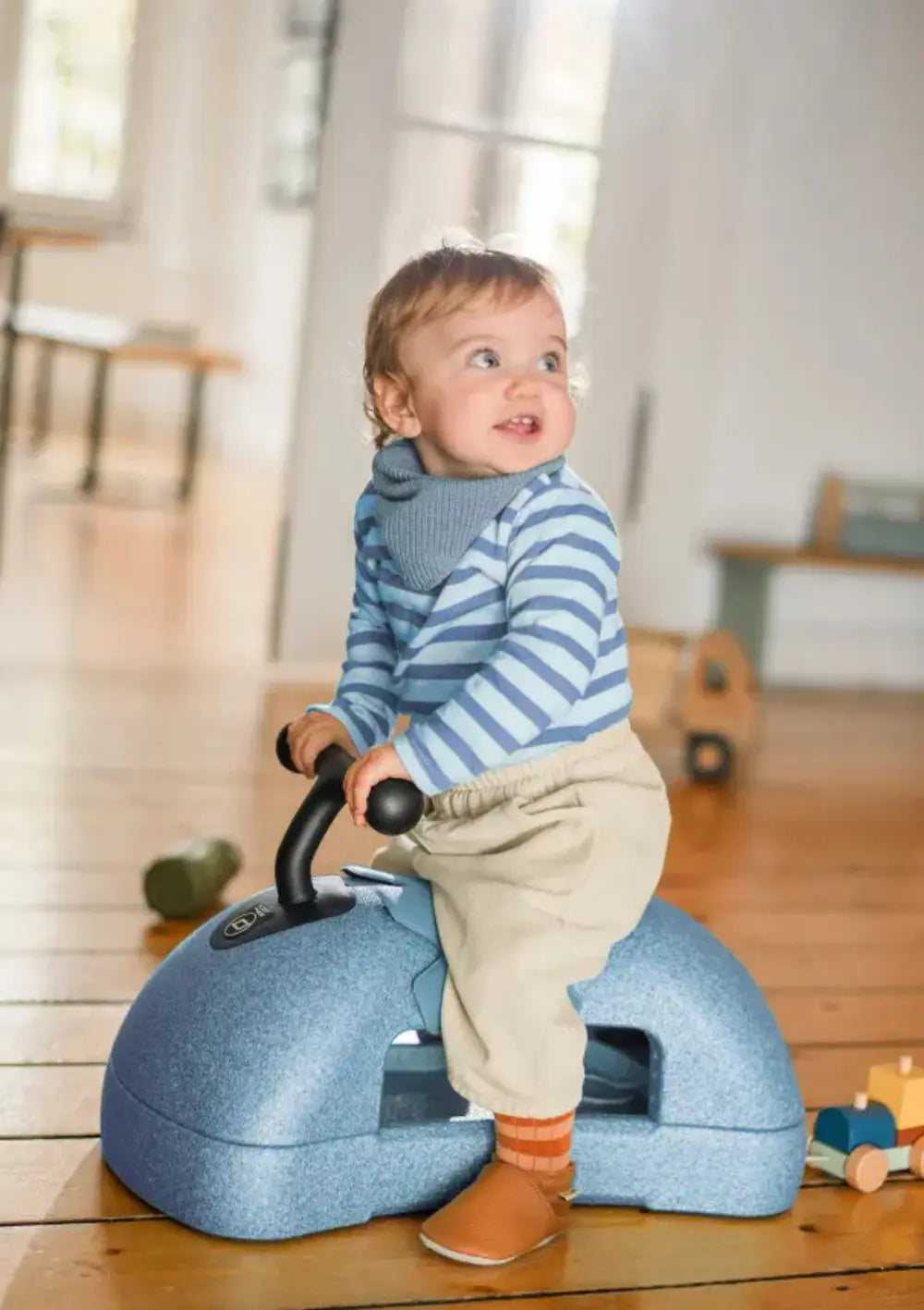 Ein Kleinkind im blau gestreiften Oberteil sitzt auf dem taubenblauen Scoot and Ride 3in1 Babyroller My First Steel und nutzt ihn in der Wohnung als komfortables Rutschfahrzeug.