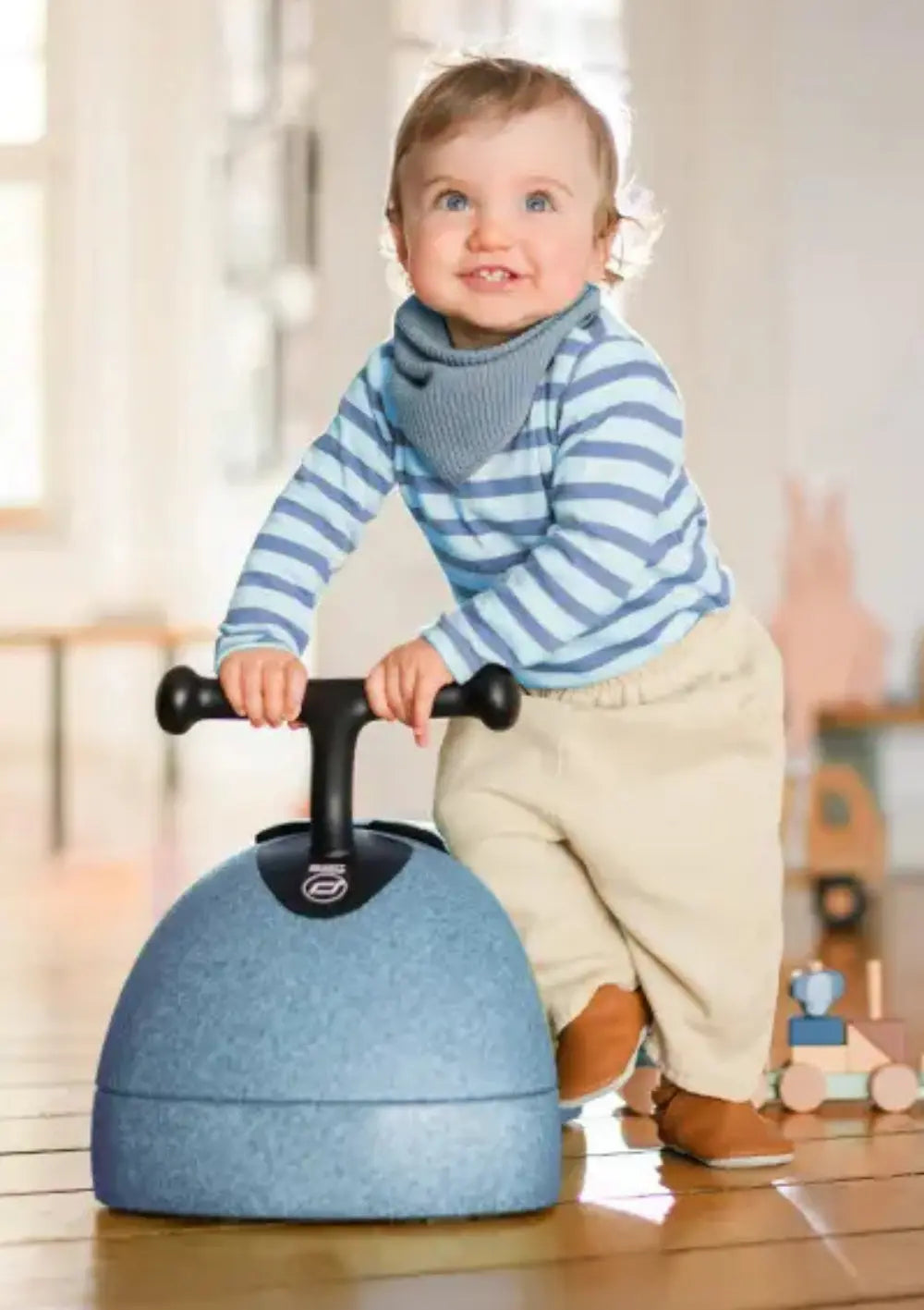 Ein lächelndes Baby steht auf dem Holzboden und hält sich am T-Lenker des blauen Scoot and Ride 3in1 Babyroller My First Steel fest, um das Gleichgewicht zu trainieren.