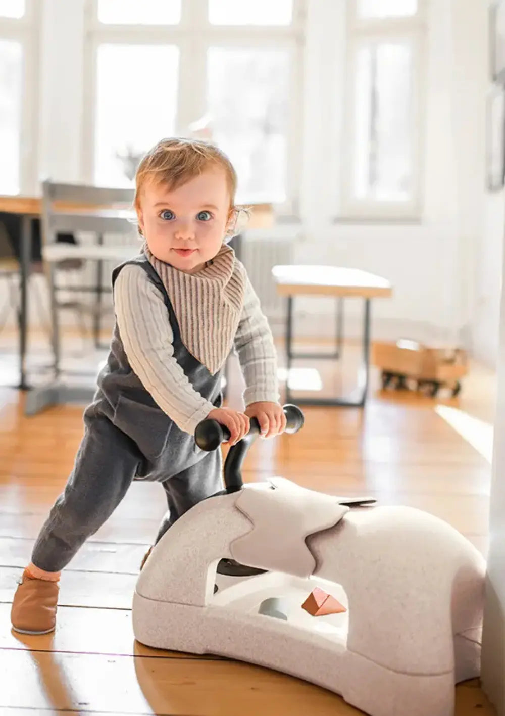 Ein Kind im grauen Strampler schiebt den beigefarbenen Scoot and Ride 3in1 Babyroller My First Sand und nutzt das multifunktionale Spielzeug als Balancier- und Lauflernhilfe im Zimmer.