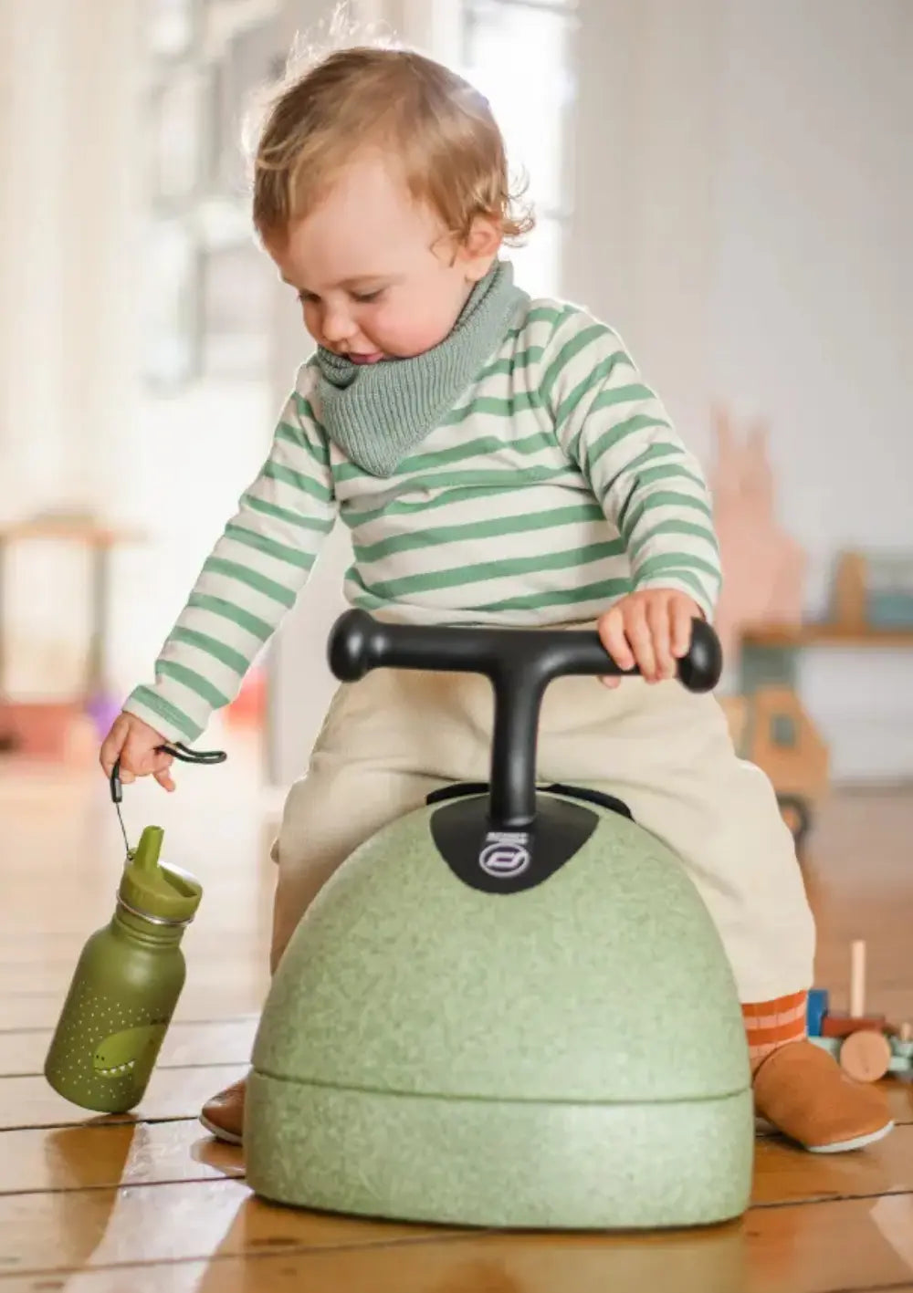 Ein gestreift gekleidetes Kind sitzt auf dem Scoot and Ride 3in1 Babyroller My First Olive und hält in einer Hand eine farblich passende, grüne Kinder-Trinkflasche.