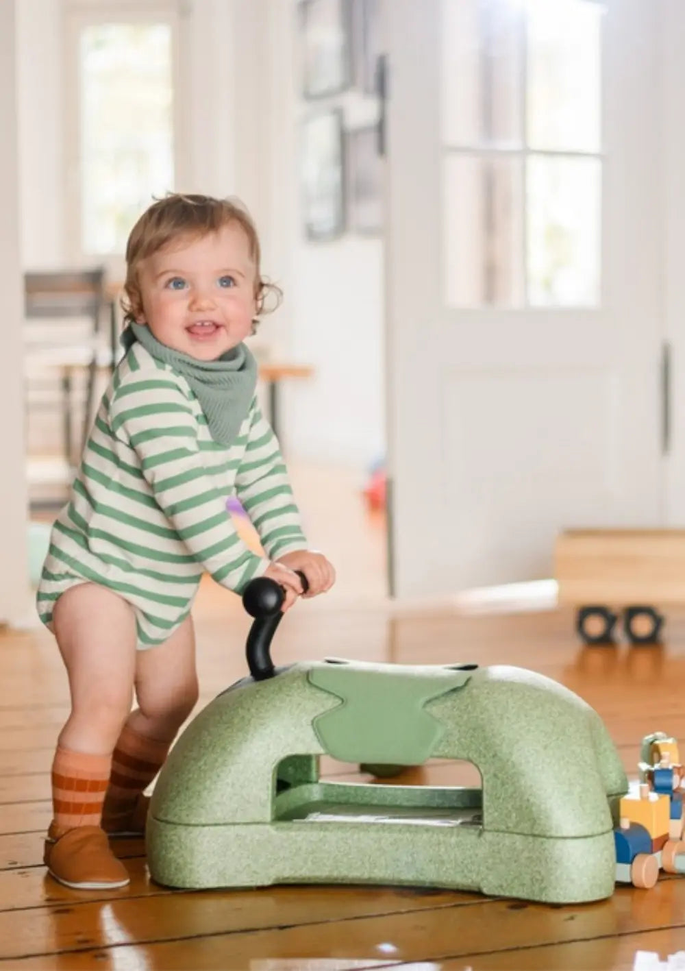 Ein fröhliches Kleinkind nutzt den olivgrünen Scoot and Ride 3in1 Babyroller My First Olive als stabile Lauflernhilfe und schiebt das ergonomische Rutschtier über den Holzboden.