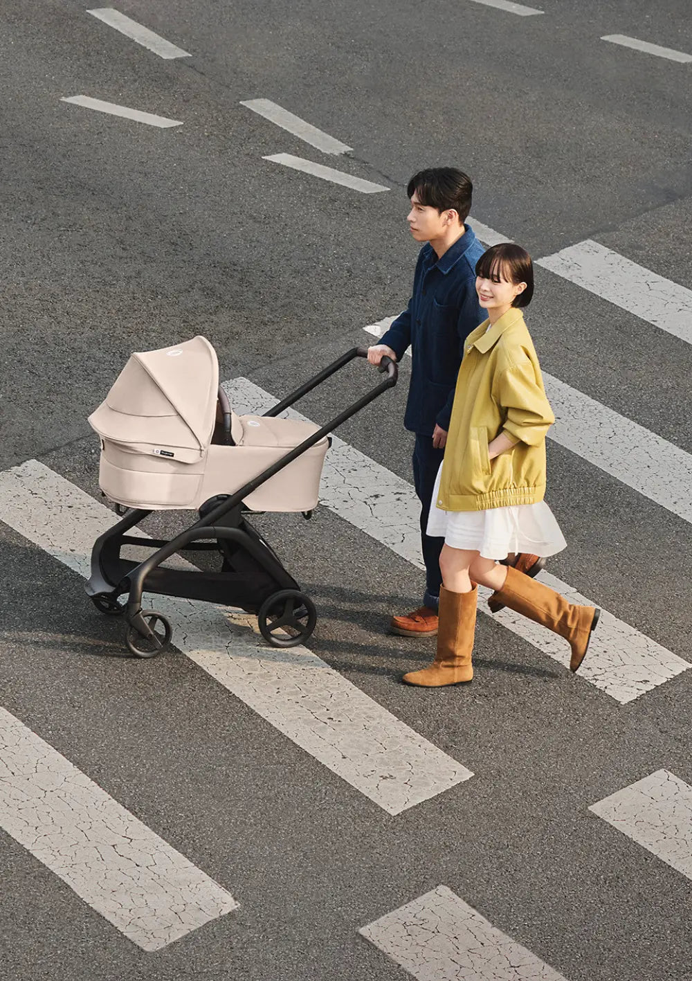 Ein junges Paar überquert einen Zebrastreifen mit dem urbanen Kombikinderwagen, auf dem die beigefarbene Bugaboo Liegewanne für den Kinderwagen Dragonfly Plus montiert ist.