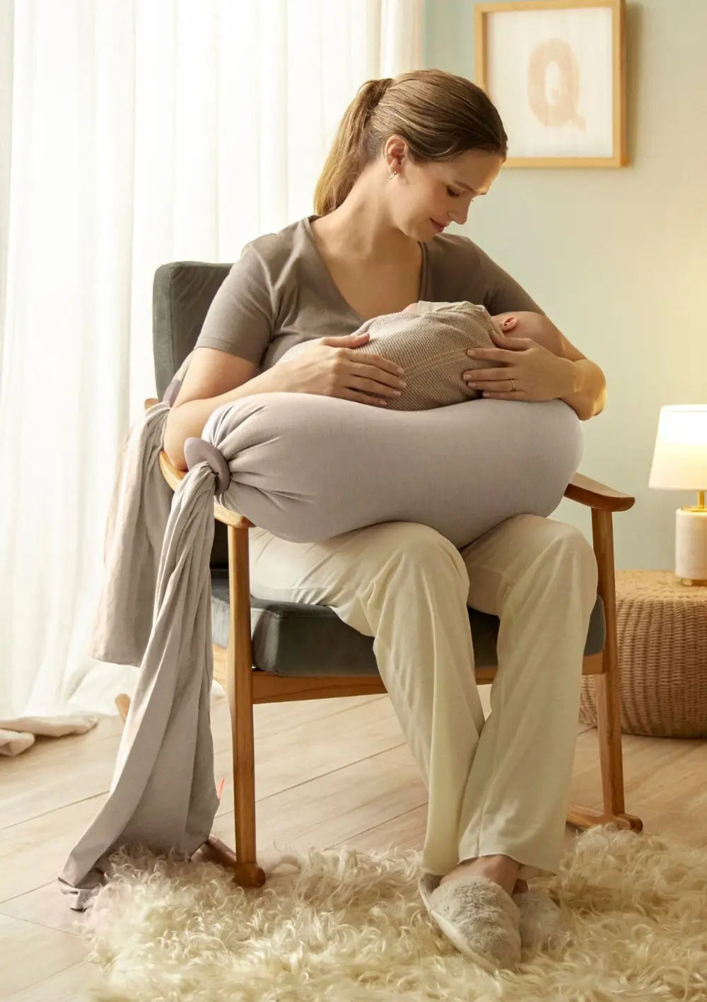 Eine Mutter sitzt in einem Sessel und nutzt das bbhugme Schwangerschaftskissen Seashell Beige u-förmig auf ihrem Schoß als entlastendes Stillkissen, um ihr schlafendes Säugling geborgen zu halten.