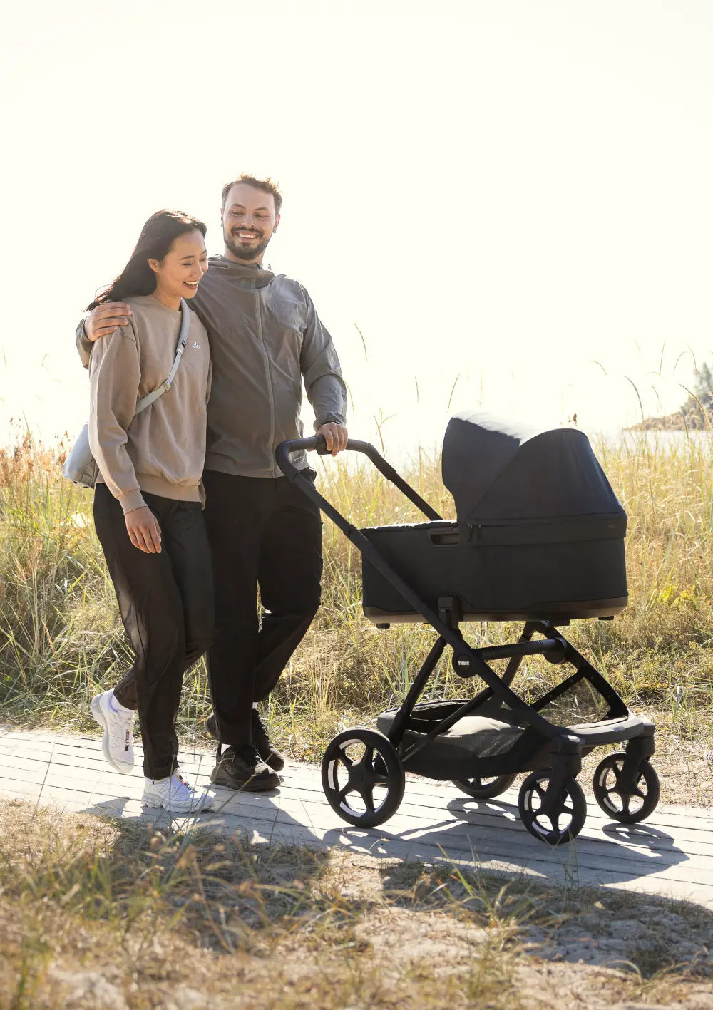 Ein lächelndes Paar spaziert bei Sonnenschein auf einem Holzsteg durch hohe Gräser mit dem Thule Kinderwagen-Set 'Charm' Black.