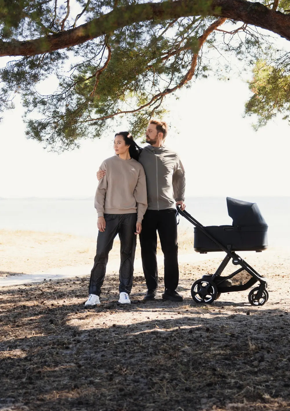 Eltern stehen mit dem Thule Kinderwagen-Set 'Charm' Black im Schatten einer großen Kiefer an einem hellen Strandabschnitt und blicken in die Ferne.