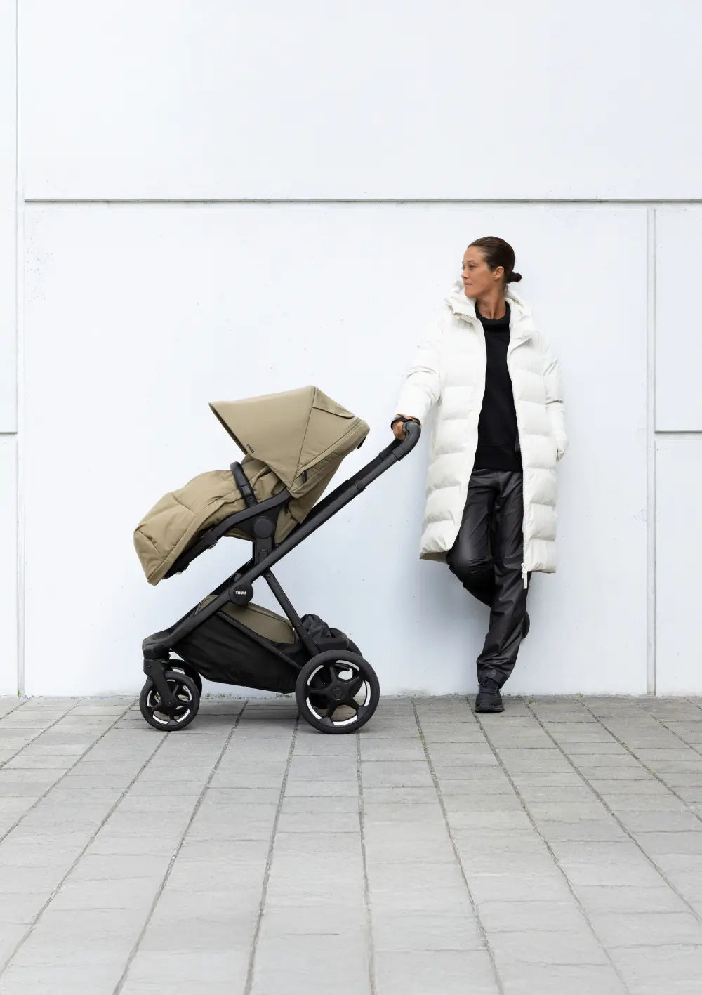 Seitenansicht des Thule Kinderwagen-Set 'Charm' Faded Khaki mit passendem Fußsack. Eine Frau lehnt im Hintergrund an einer hellen Fassade und blickt zum Kinderwagen.