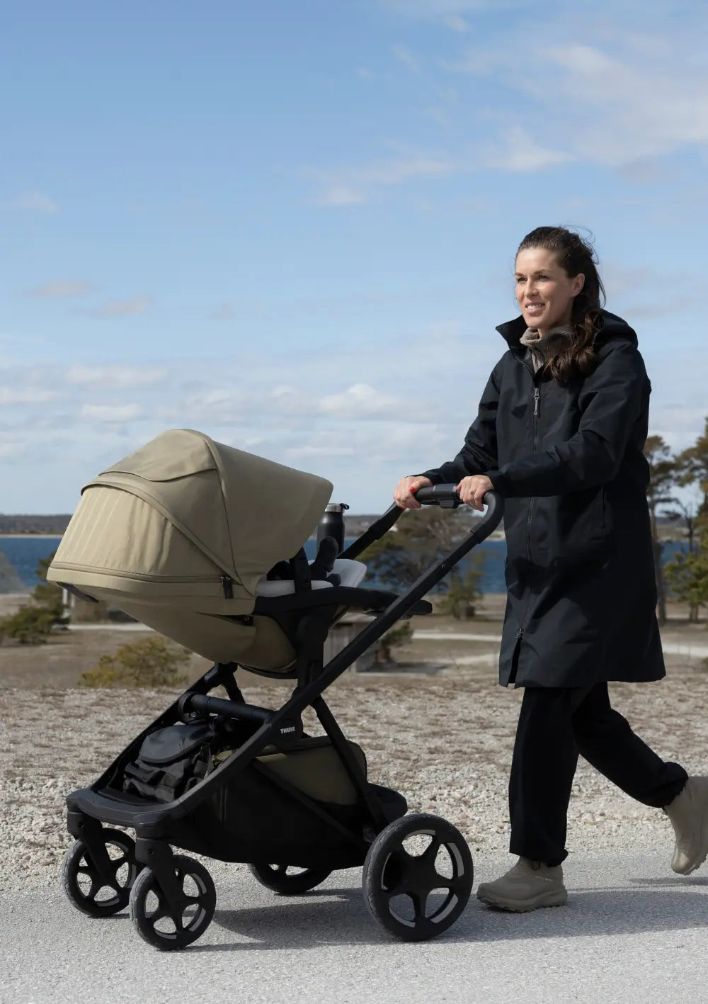 Eine Frau schiebt das Thule Kinderwagen 'Charm' Faded Khaki mit Sportsitz auf einem befestigten Weg in einer Küstenlandschaft unter blauem Himmel.