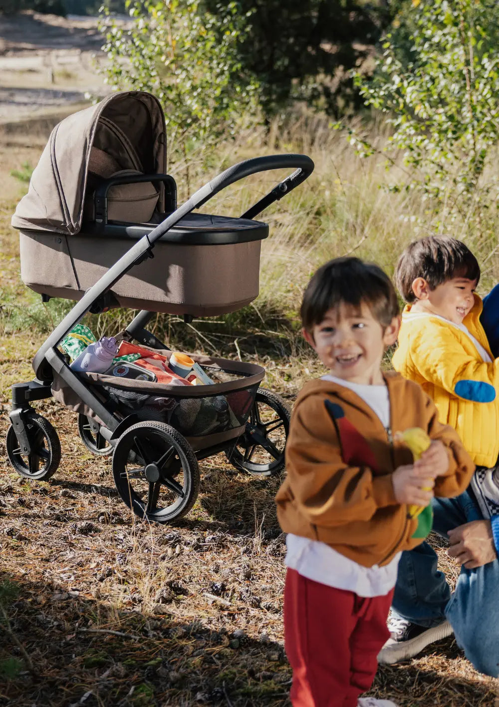 Zwei Kinder spielen in der Natur – im Hintergrund: Joolz Kinderwagen-Set 'Geo5' Single Hazel Brown mit vollbepacktem Staukorb