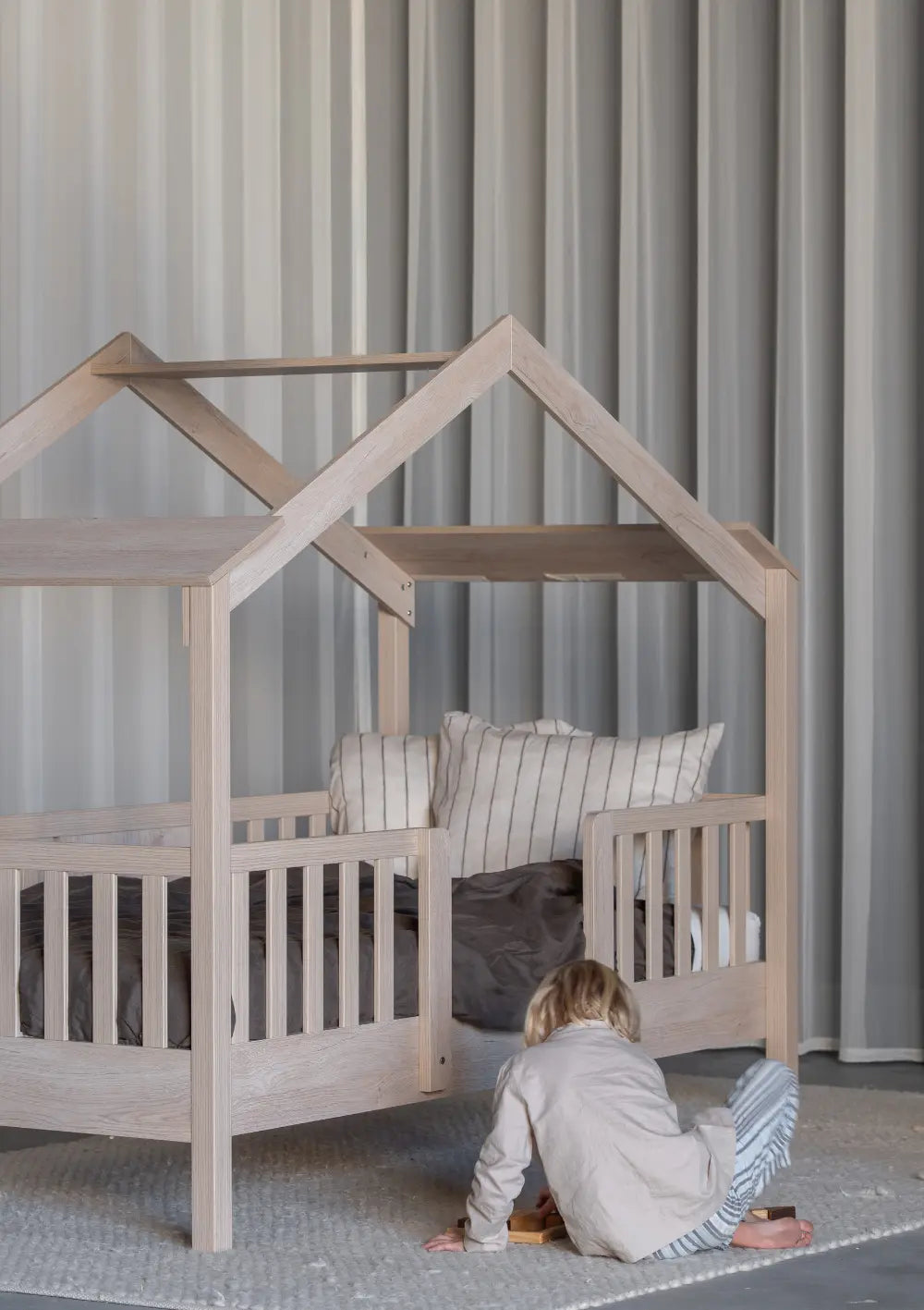 Ein Kind spielt auf dem Teppich vor dem Quax Hausbett Chalet Natural Ash (200 x 90 cm); das Bettgestell aus hellem Holz in Haus-Optik verfügt über einen integrierten Rausfallschutz und ein offenes Dachgestell.