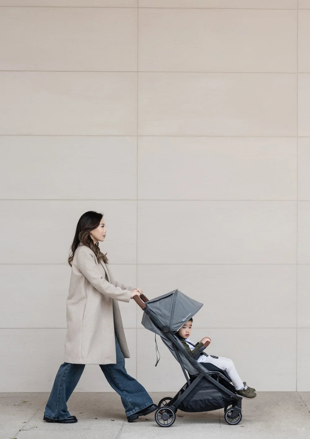 Eine Frau im beigen Mantel und blauer Jeans schiebt ein Kleinkind im UPPAbaby Buggy Minu V3 Greyson vor einer minimalistischen, hellen Wand.