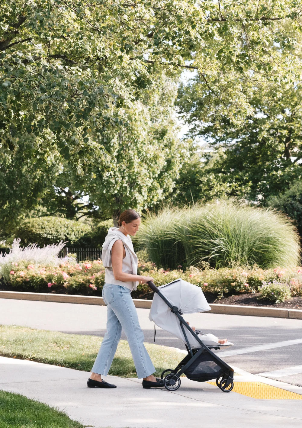 Komfortabler City-Buggy: Eine Frau schiebt den UPPAbaby Buggy Minu V3 Savannah in der Seitenansicht über einen gepflasterten Gehweg vorbei an einer gepflegten Parkanlage.