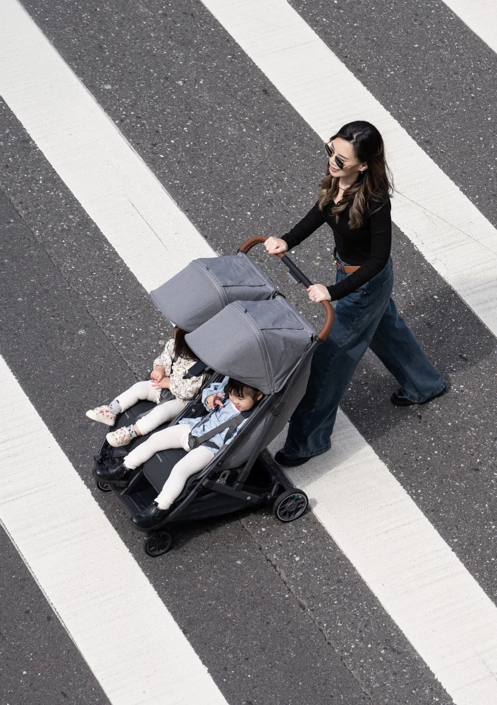 Blick von oben: Eine Frau überquert mit dem UPPAbaby Geschwisterbuggy Minu Duo Greyson einen Zebrastreifen; zwei Kinder sitzen nebeneinander im Buggy.