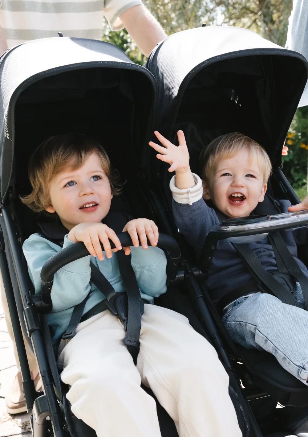 Aufnahme von zwei Kleinkindern, die nebeneinander im UPPAbaby Geschwisterbuggy Minu Duo Jake sitzen, lachen und winken.