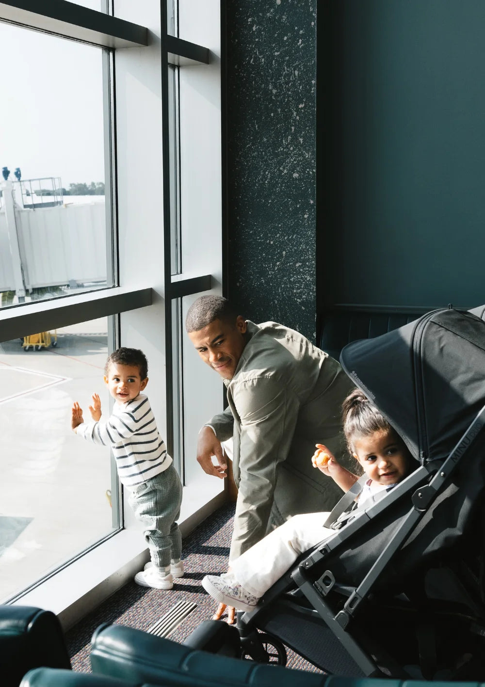 Reisebereit mit Doppel-Buggy: Ein Vater und zwei Kinder im Flughafen-Wartebereich; ein Kind sitzt im UPPAbaby Geschwisterbuggy Minu Duo Jake.