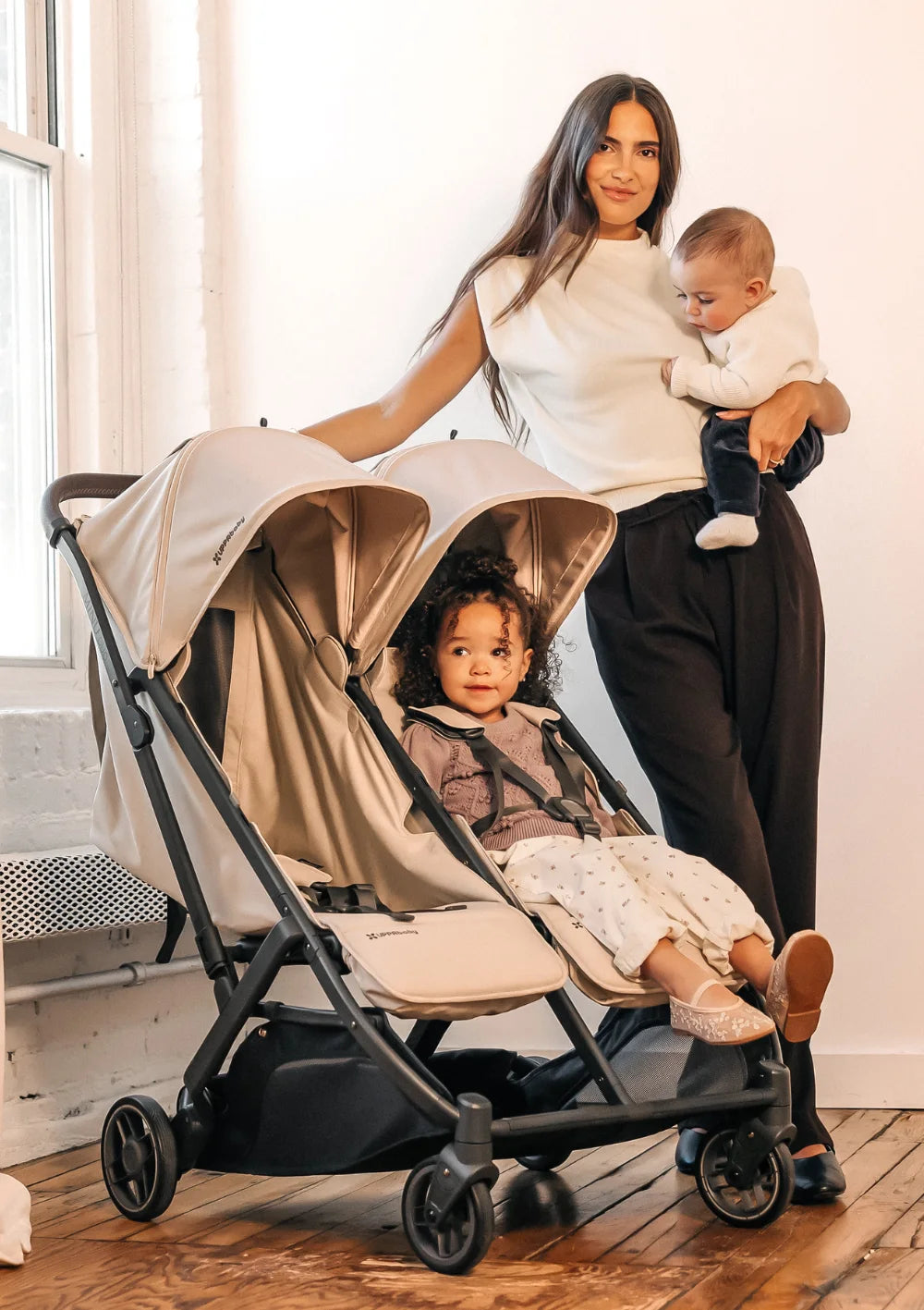 Eine Mutter mit Baby auf dem Arm steht neben dem UPPAbaby Geschwisterbuggy Minu Duo Ada, in dem ein weiteres Kind sitzt, in einem hellen Raum mit Holzfußboden.
