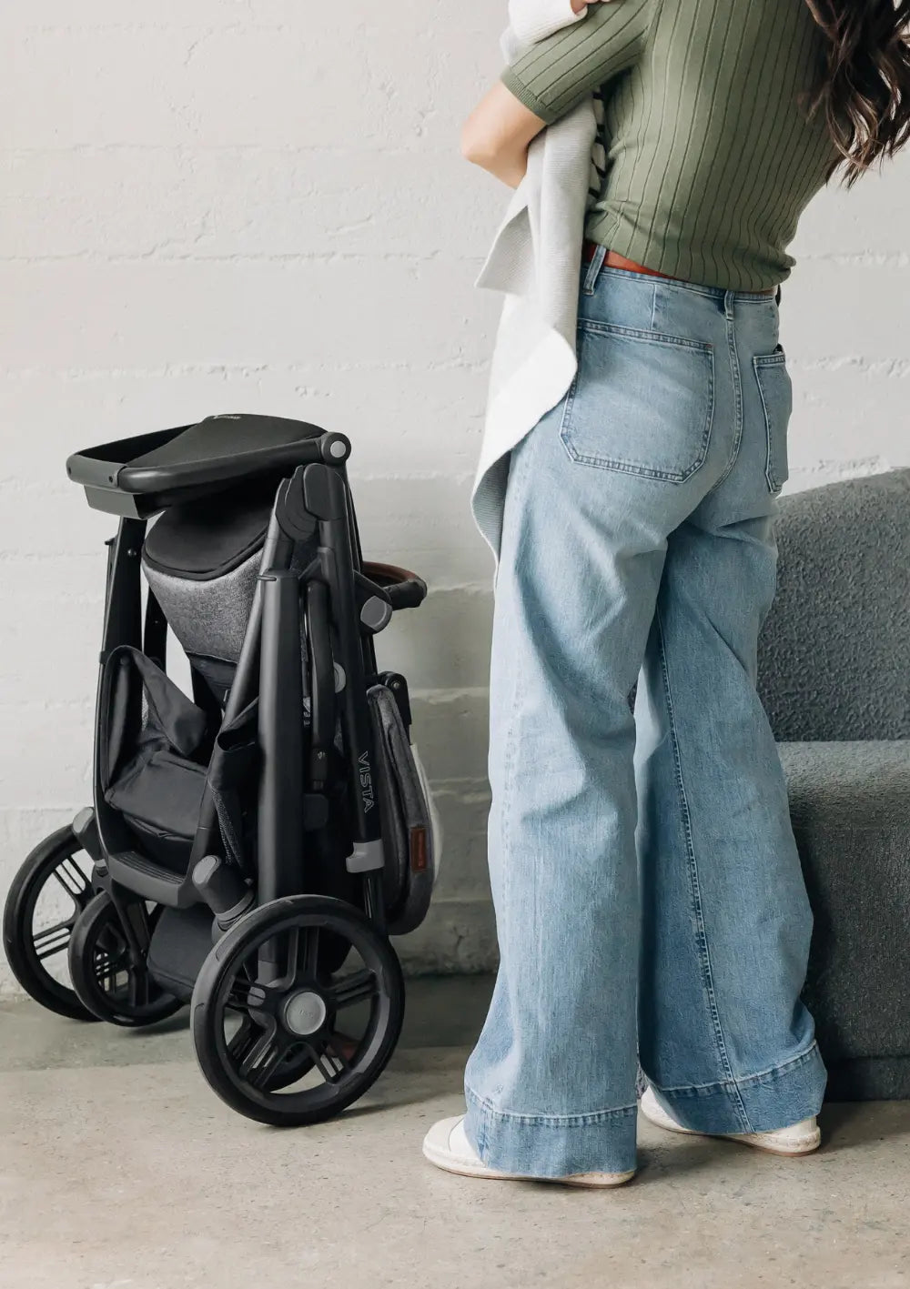 Eine Frau hält ihr Kind im Arm; daneben steht der zusammengeklappte, selbststehende UPPAbaby Kinderwagen 'Vista V3' Greyson