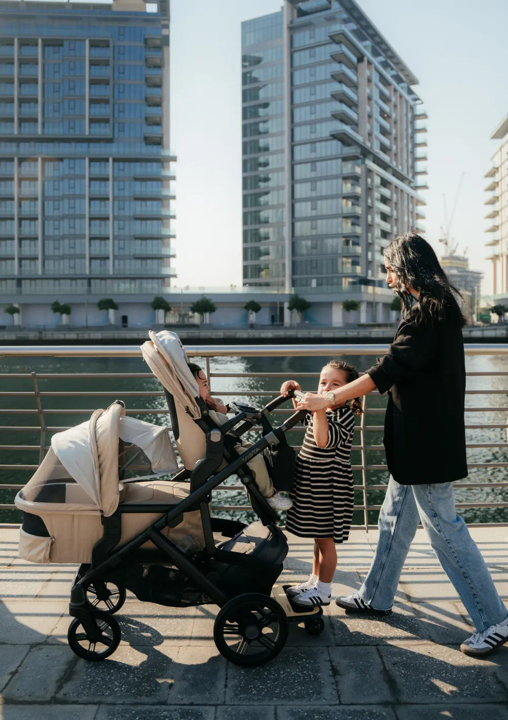 Eine Frau ist mit drei Kindern an einer Uferpromenade in der Stadt unterwegs. Im Bild: das Kinderwagen-Set UPPAbaby 'Vista 3' Liam mit Babywanne, Sportsitz und Mitfahrbrett