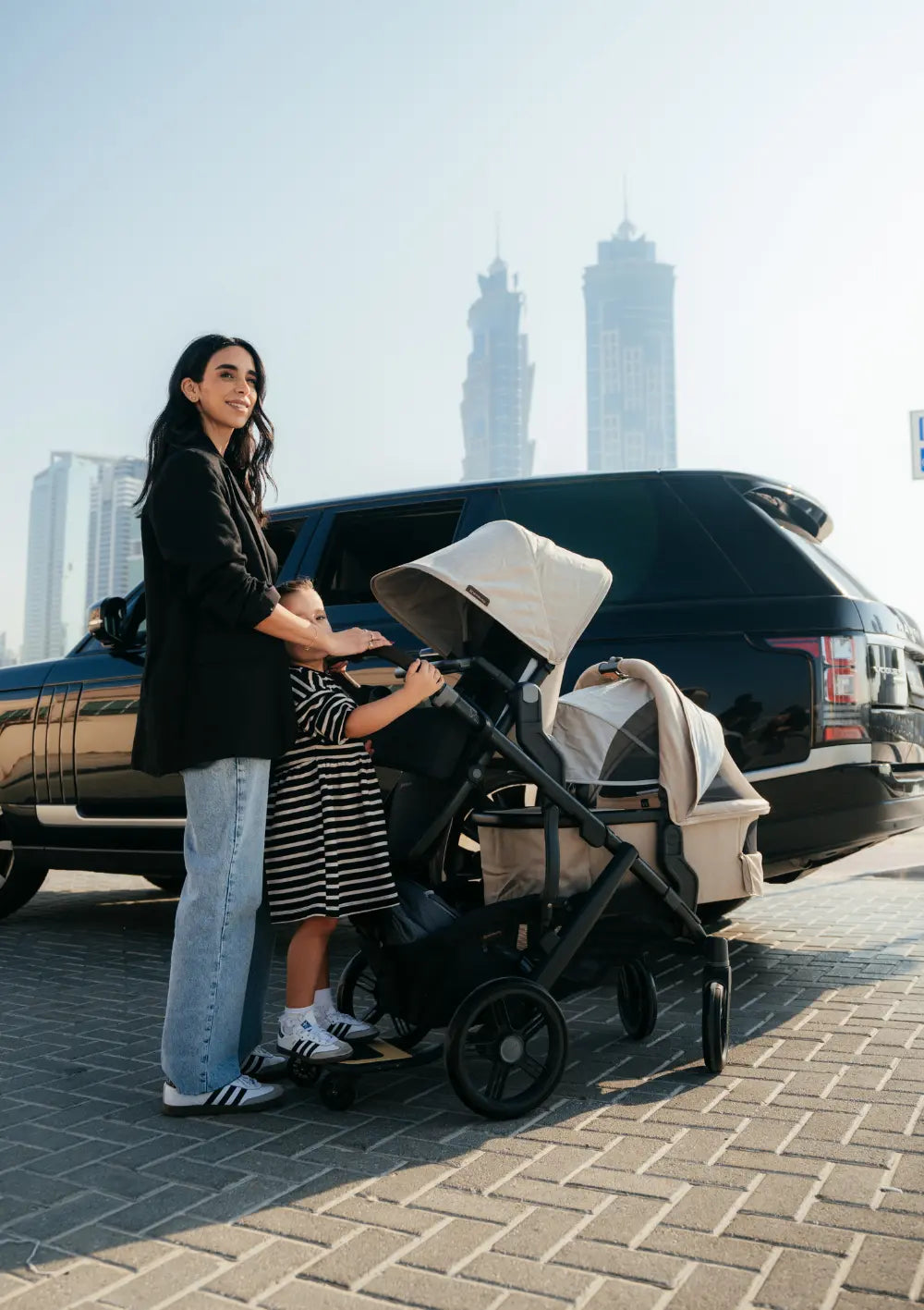 Frau steht mit Kindern im Kinderwagen-Set UPPAbaby 'Vista 3' Liam mit Babywanne, Sportsitz und Mitfahrbrett vor einem Auto