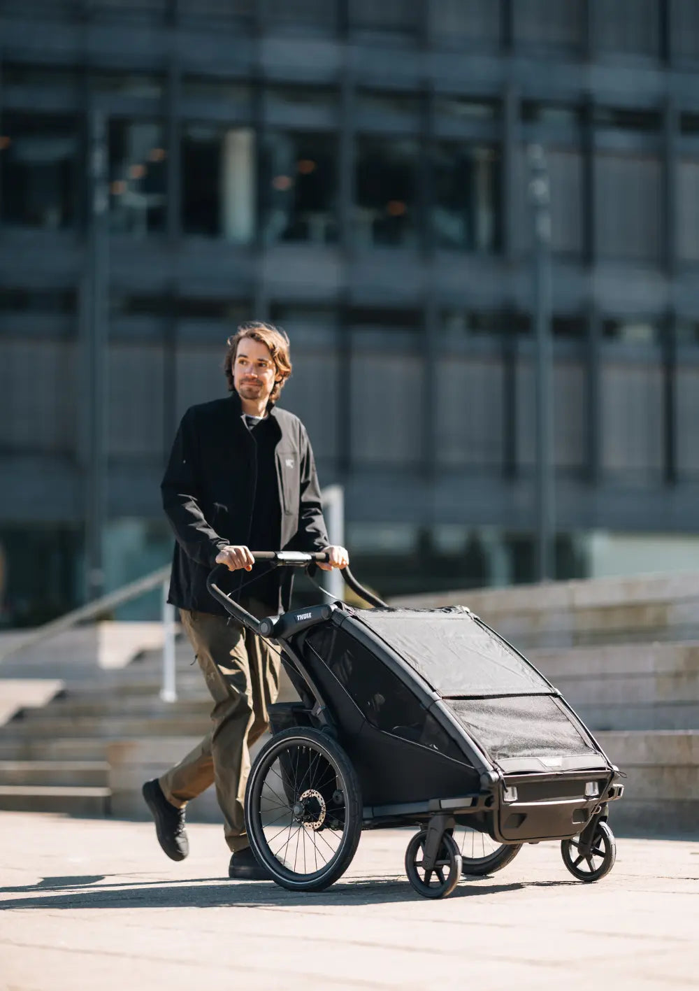 Urbaner Einsatz: Ein Mann schiebt den Thule Multisport-Fahrradanhänger Chariot Sport 2 Double Black im Buggy-Modus vor einem modernen Bürogebäude; flexibler Zweisitzer für die Stadt.