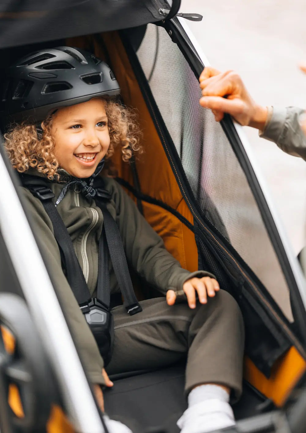 Ein lächelndes Kind mit Locken und Fahrradhelm sitzt sicher angeschnallt im Thule Multisport-Fahrradanhänger Chariot Sport 2 Single Natural Gold.