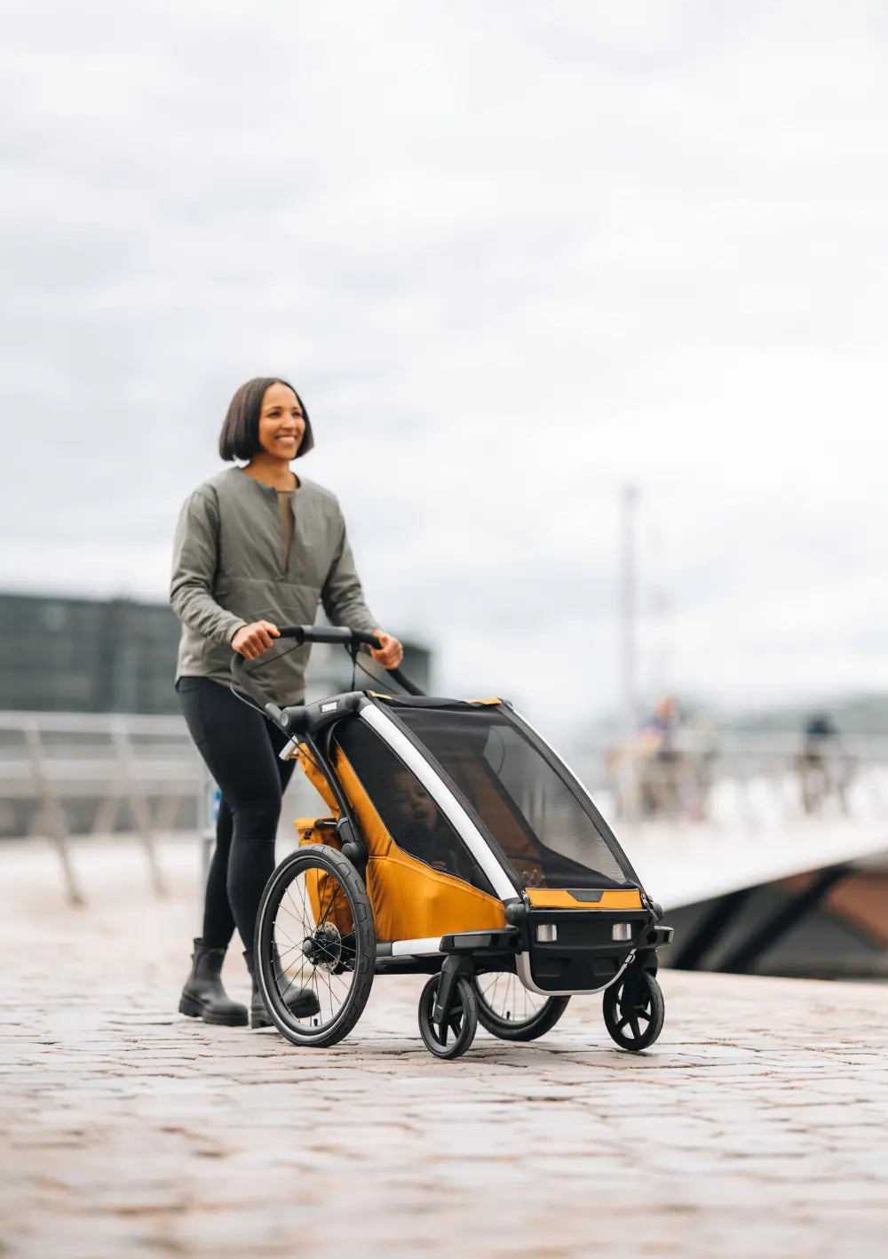 Eine Frau schiebt den Thule Multisport-Fahrradanhänger Chariot Sport 2 Single Natural Gold im Buggy-Modus über einen gepflasterten Weg am Wasser.