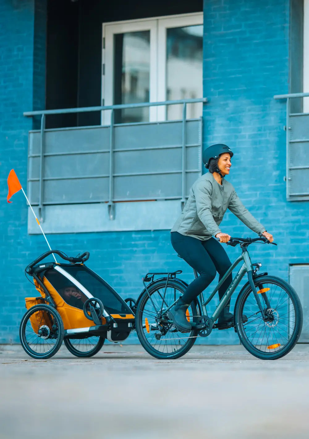 Sportlicher Lifestyle: Eine Frau fährt ein E-Bike mit dem angekoppelten Thule Multisport-Fahrradanhänger Chariot Sport 2 Single Natural Gold vor einer blauen Fassade.