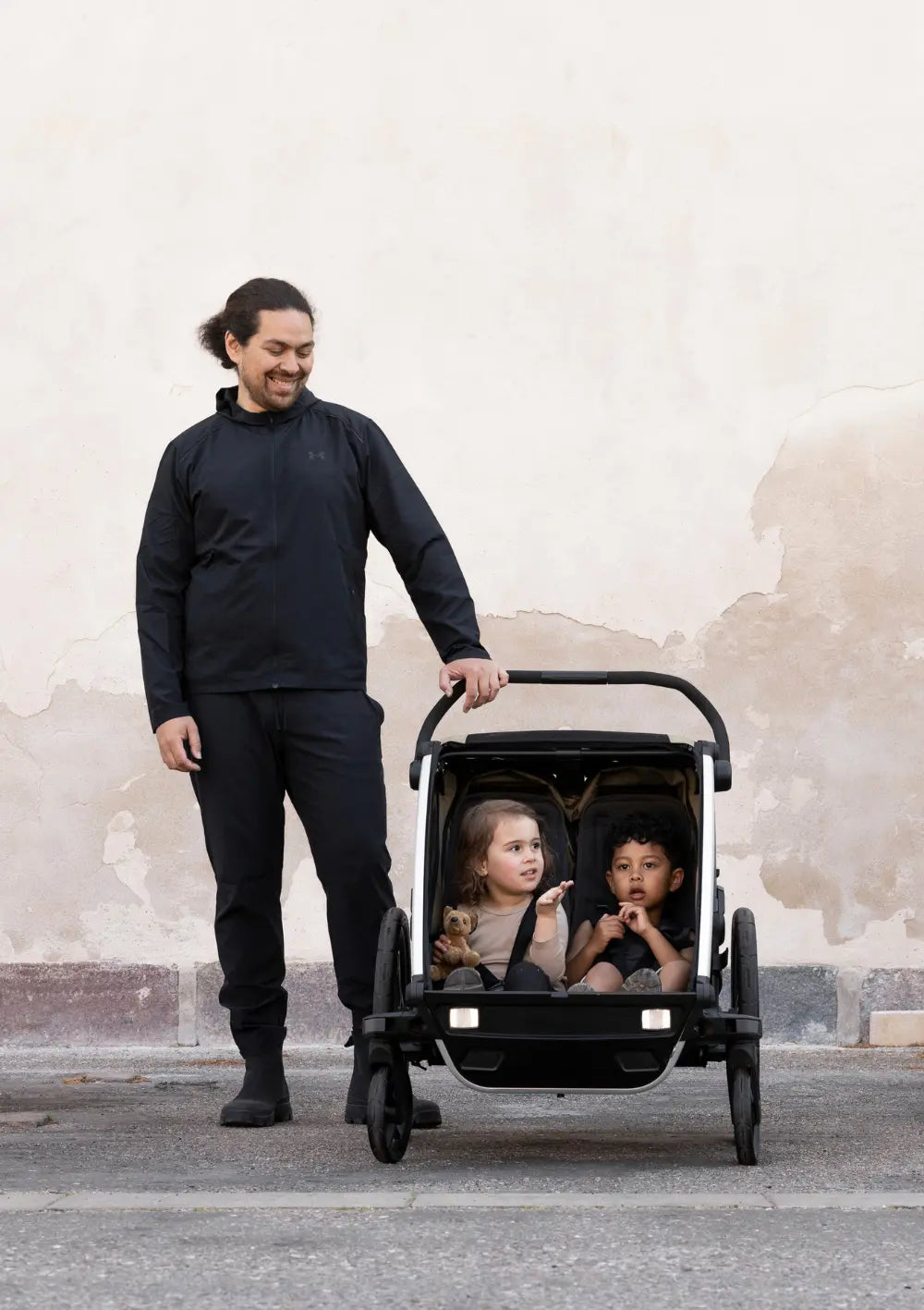 Ein Vater steht neben dem Thule Multisport-Fahrradanhänger Chariot Cross 2 Double Faded Khaki, in dem zwei Kinder nebeneinander sitzen; Fokus auf die Zweisitzer-Funktion vor einer hellen Wand.