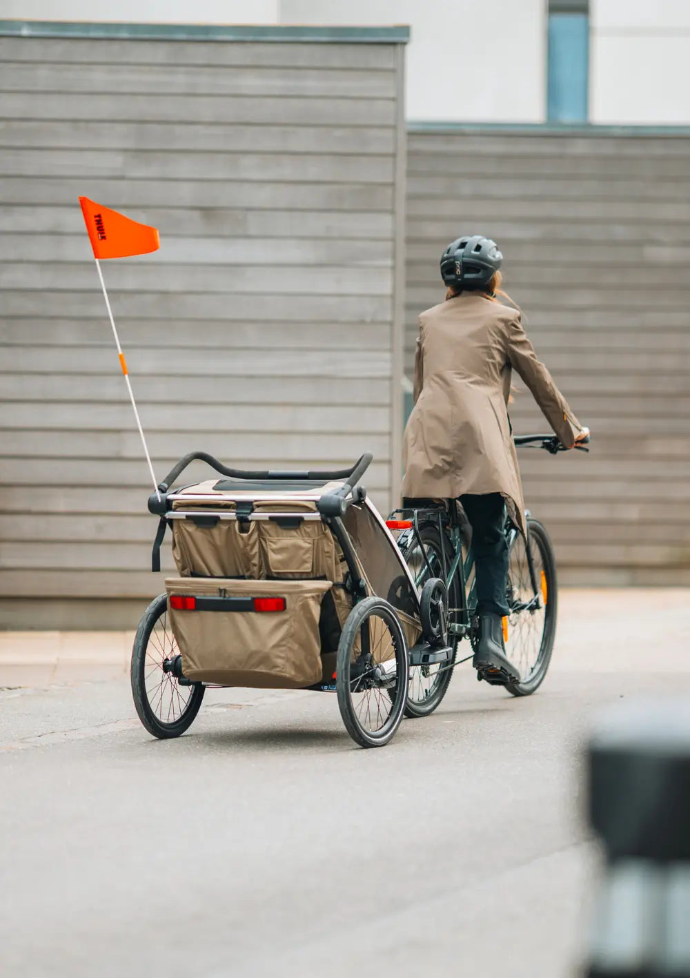 Rückansicht einer Frau beim Radfahren mit dem angekoppelten Thule Multisport-Fahrradanhänger Chariot Cross 2 Double Faded Khaki mit Sicherheitswimpel und großen Reflektoren.