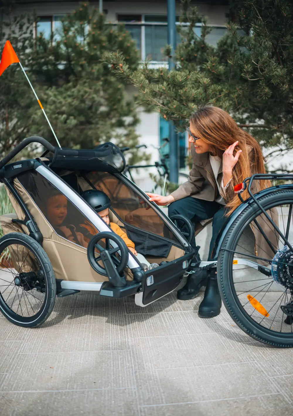 Eine Mutter schaut lächelnd zu ihren beiden Kindern im Thule Multisport-Fahrradanhänger Chariot Cross 2 Double Faded Khaki.