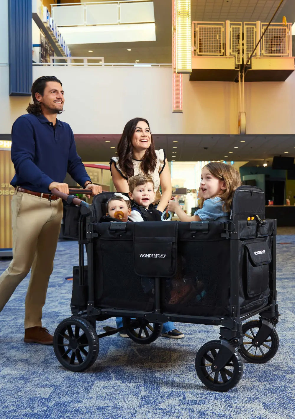 Eine lächelnde Familie mit drei Kindern nutzt den WonderFold W4 Luxe Pro Kinderwagen 4-Sitzer Jet Schwarz in einem modern gestalteten Innenbereich.