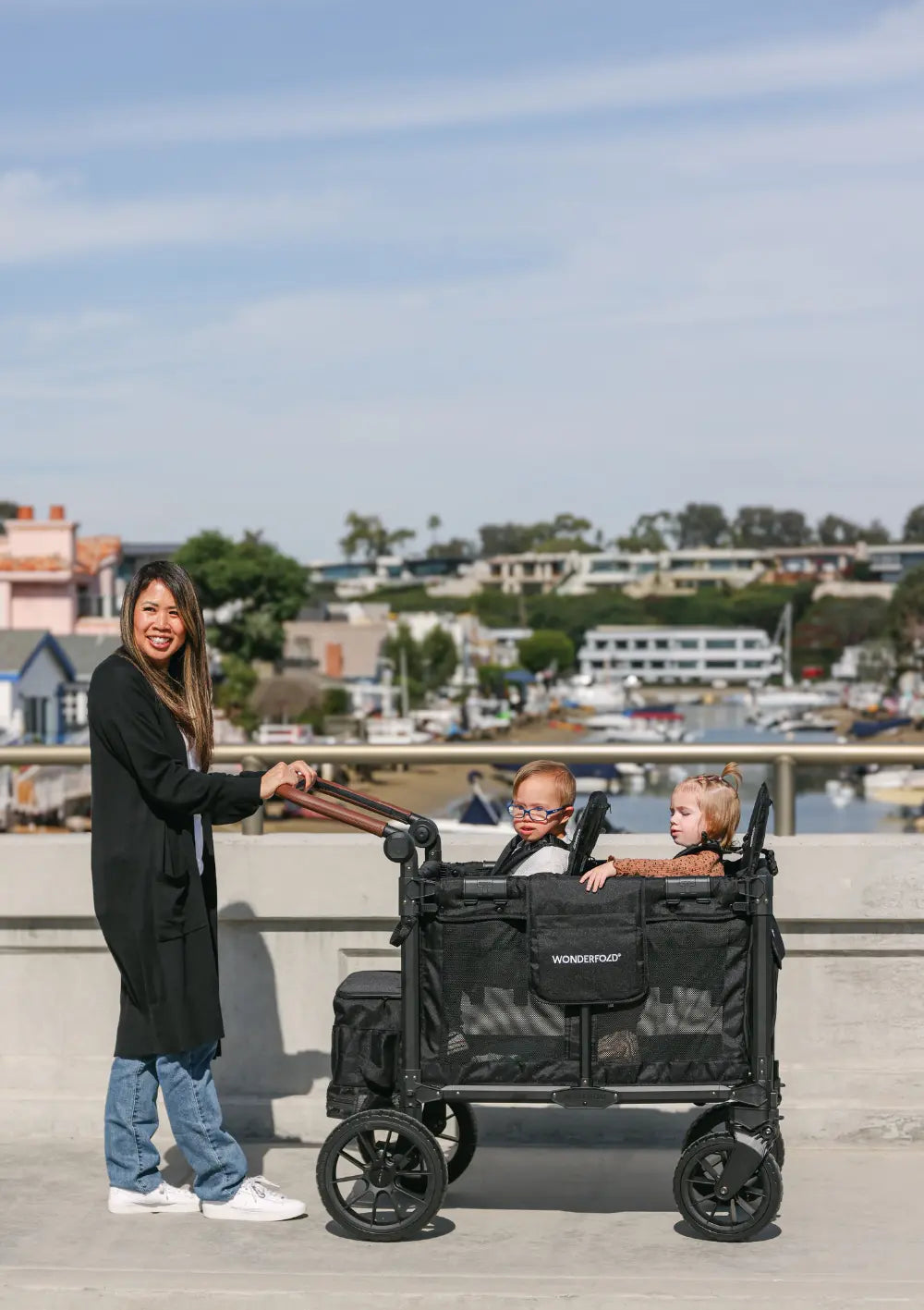 Eine Frau schiebt den WonderFold W2 Luxe Pro Kinderwagen 2-Sitzer Jet Schwarz über eine sonnige Brücke mit Blick auf eine Marina im Hintergrund.