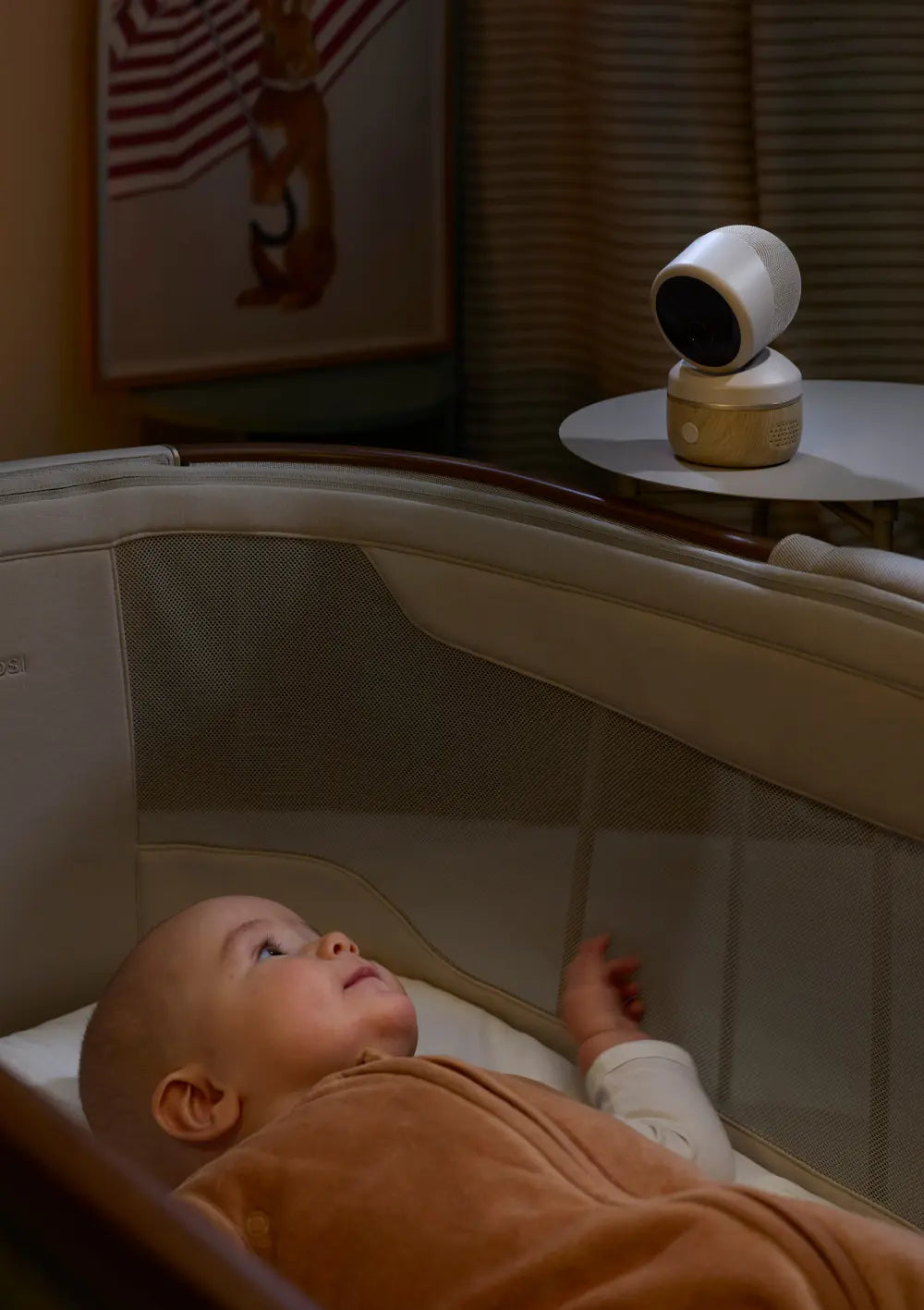 Das smarte Maxi-Cosi Babyphone Connected Home See 2 Plus steht auf einem runden Tischchen neben dem Beistellbett, während das Baby wach darin liegt.