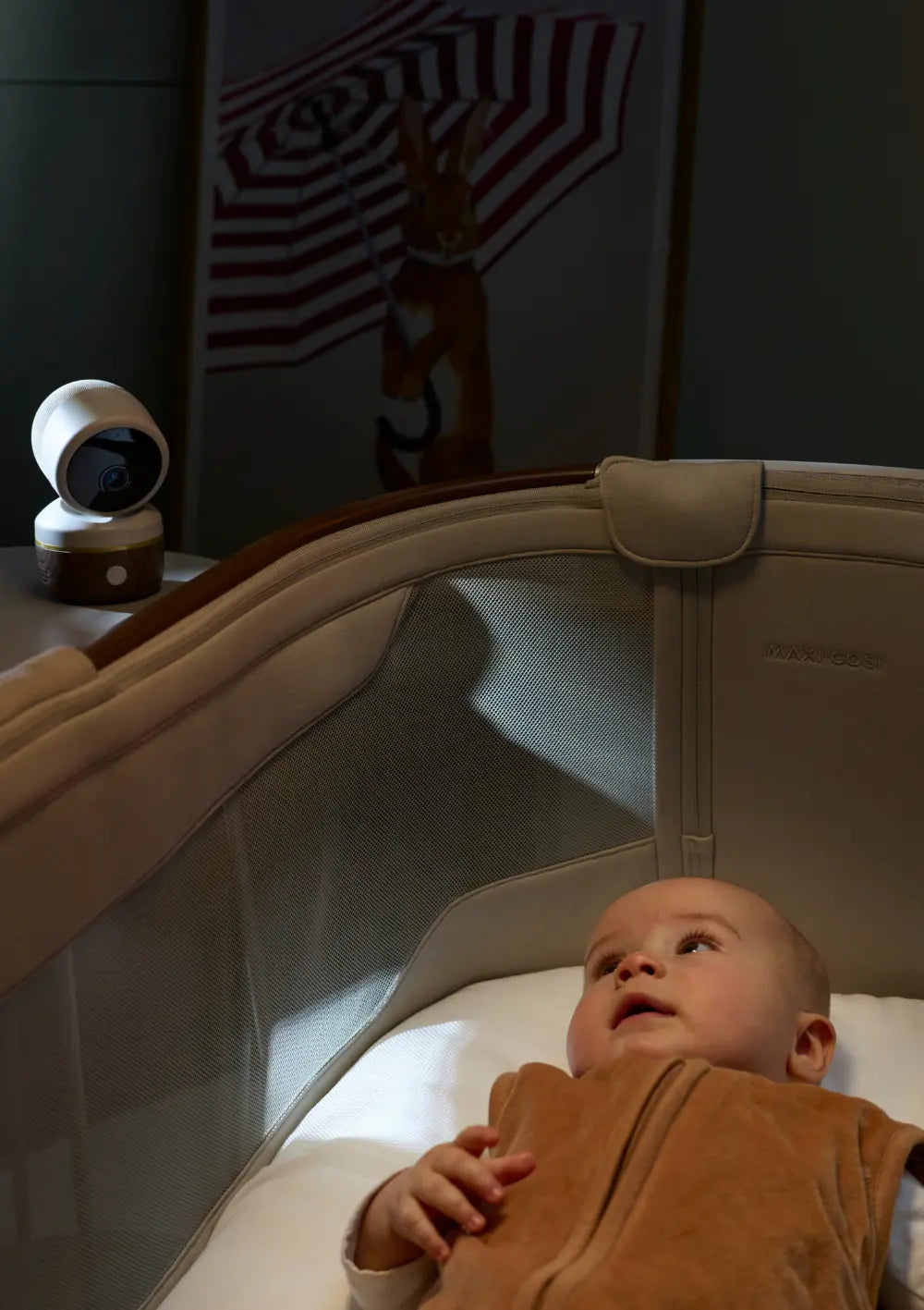 Der Maxi-Cosi Smart Baby Monitor Connected Home See 2 Pro Elegance Beige steht auf einem Tischchen neben einem Babybett und filmt das darin liegende, wach nach oben schauende Baby.