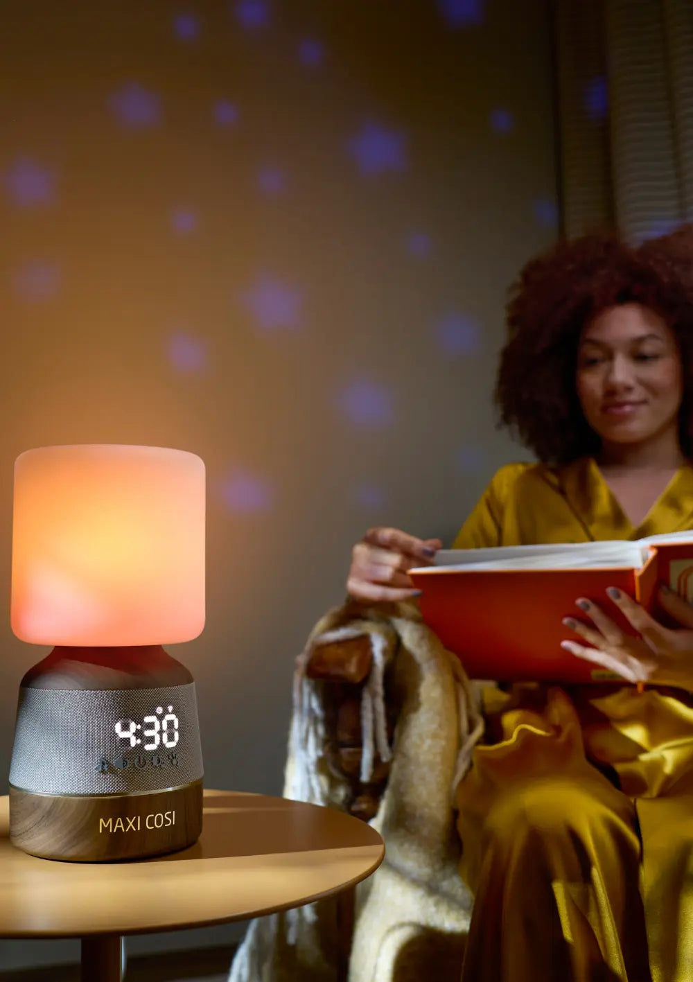 Das Maxi-Cosi Nachtlicht & Klang Connected Home Soothe Pro wirft eine Sternenprojektion an die Wand, während eine Frau daneben im warmen Schein der Lampe ein Buch liest.