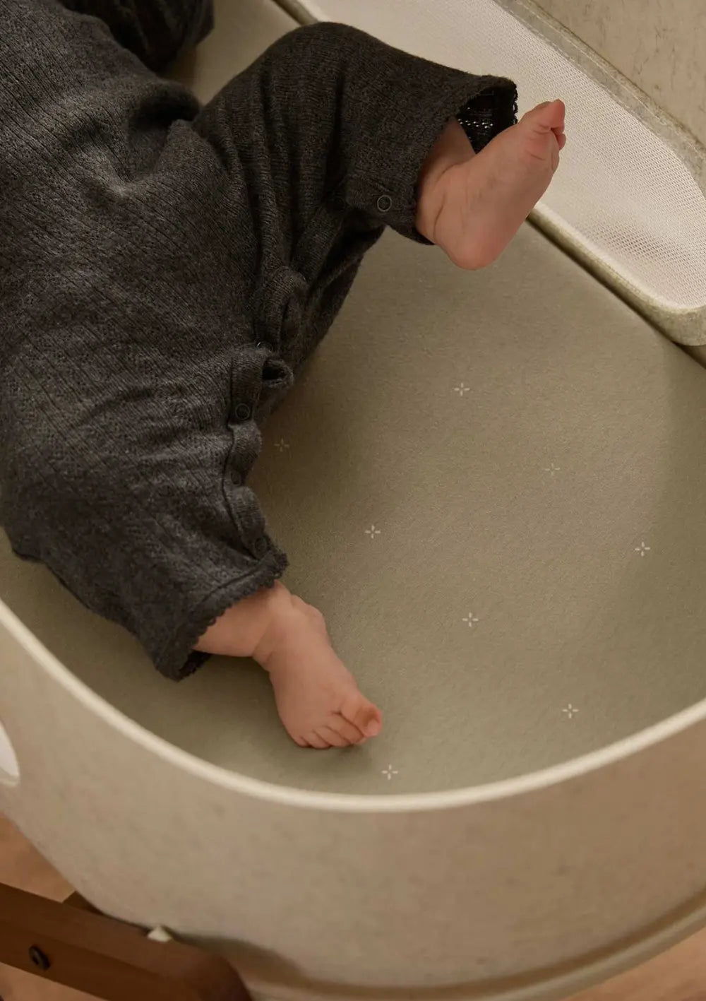 Nahaufnahme der Füße eines Babys im dunklen Strampler auf dem salbeigrünen Snüz Spannbettlaken für Moseskorb SnüzBaskit Sage mit Kreuzchen.