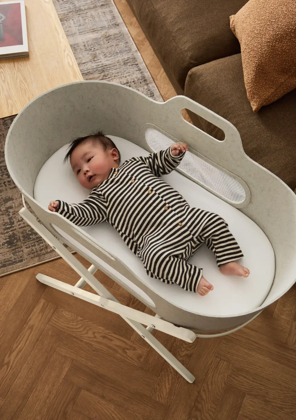 Ein auf dem Rücken liegendes Baby im gestreiften Strampler blickt neugierig aus dem hellbeigen Snüz Moseskorb inkl. Ständer SnüzBaskit Barley-Cashmere; Draufsicht auf den gemütlichen Schlafplatz.
