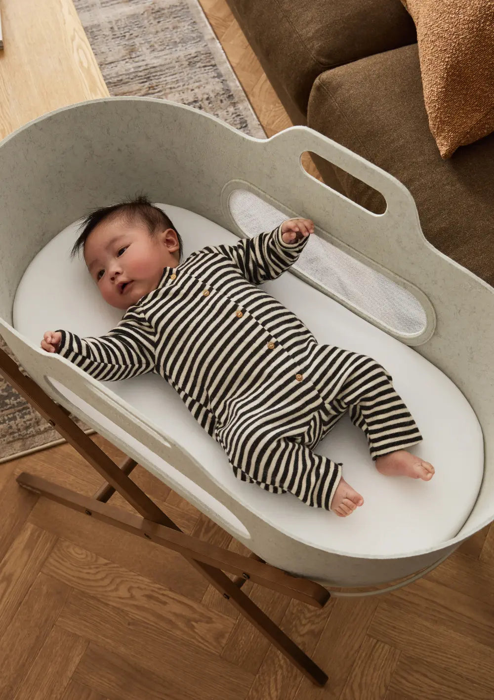 Blick von oben auf ein liegendes Baby im hellbeigen Snüz Moseskorb inkl. Ständer SnüzBaskit Barley-Walnuss mit komfortabler, weißer Matratze.