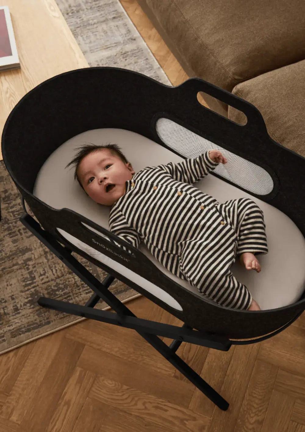 Ein Baby im gestreiften Einteiler liegt entspannt im dunkelgrauen Snüz Moseskorb inkl. Ständer SnüzBaskit Graphite; Draufsicht auf die gemütliche Babywiege.
