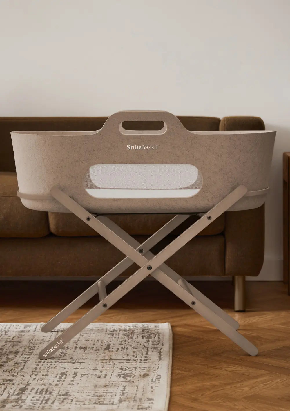 Der stilvolle Snüz Moseskorb inkl. Ständer SnüzBaskit Mocha steht komplett aufgebaut auf seinem klappbaren, grauen Holzgestell im modern eingerichteten Wohnzimmer vor einem Sofa.