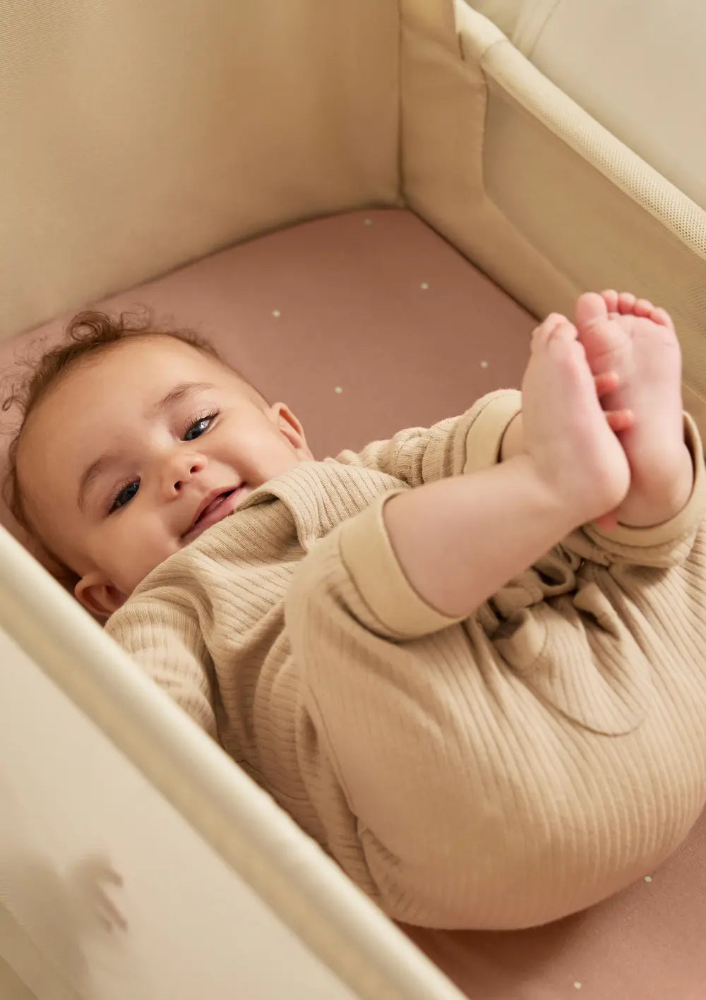 Ein lächelndes Baby liegt im cremefarbenen Snüz Beistellbett SnüzPod 5 Cashmere auf einer rosa Matratze und greift nach seinen Füßen; gemütliche Nahaufnahme.