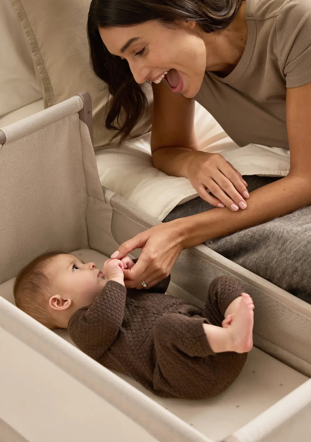 Eine Mutter liegt lächelnd im Elternbett direkt neben dem geöffneten Snüz Beistellbett SnüzPod 5 Mocha und hält die Händchen ihres Babys beim Aufwachen.