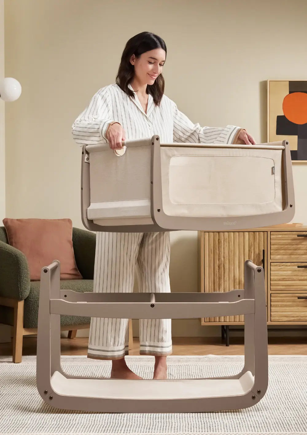 Eine Frau im gestreiften Pyjama demonstriert den flexiblen Aufbau des Snüz Beistellbettes SnüzPod 5 Mocha, indem sie die obere Liegewanne vom Holzgestell hebt.
