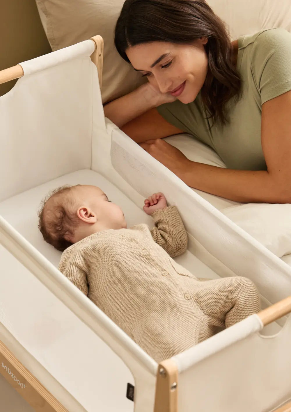 Eine Mutter blickt liebevoll auf ihr schlafendes Kleinkind, das direkt neben ihr im geöffneten Snüz Beistellbett SnüzPod 5 Natur liegt.