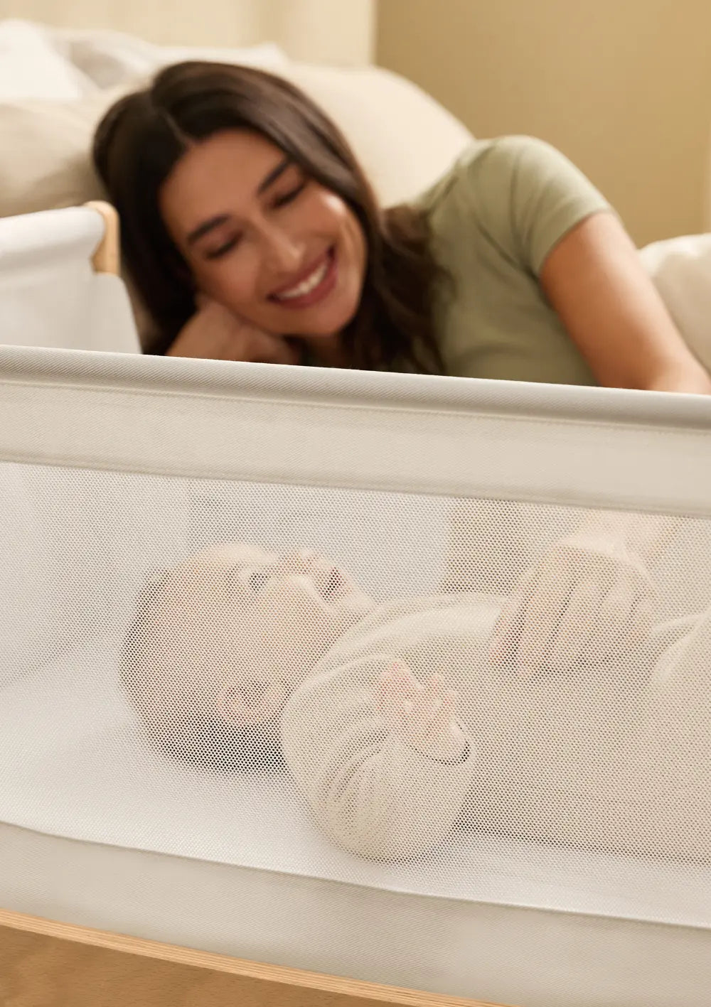 Eine lächelnde Mutter schaut von oben durch das transparente Mesh-Sichtfenster des Snüz Beistellbettes SnüzPod 5 Natur auf ihr liegendes Baby.