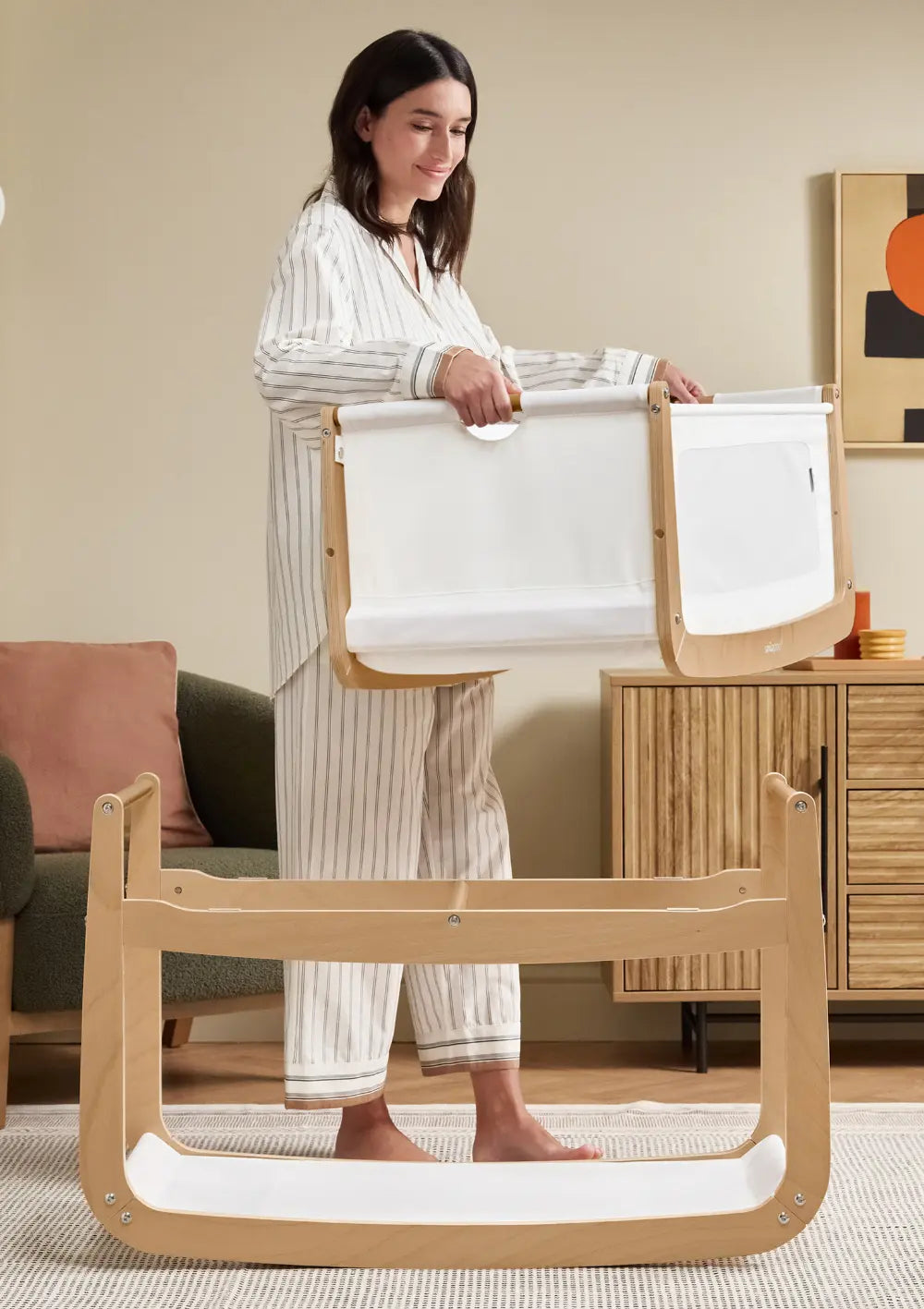 Eine Frau im gestreiften Pyjama nimmt die obere Stoff-Liegewanne des Snüz Beistellbettes SnüzPod 5 Natur vom hellen Holzgestell ab.