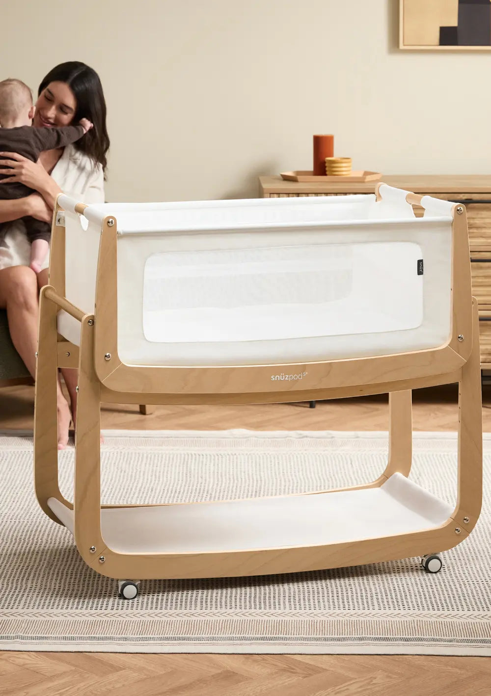 Das freistehende Snüz Beistellbett SnüzPod 5 Natur aus hellem Holz mit weißem Stoffbezug im Wohnzimmer; im Hintergrund sitzt die Mutter mit ihrem Baby.