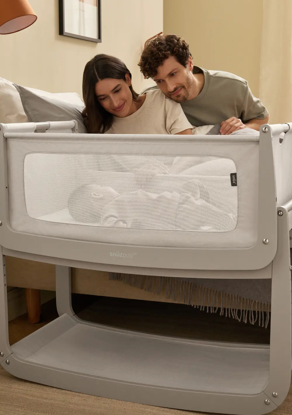 Eltern blicken gemeinsam im Schlafzimmer durch das große Mesh-Sichtfenster des grauen Snüz Beistellbettes SnüzPod 5 Pebble auf ihr liegendes Kind.