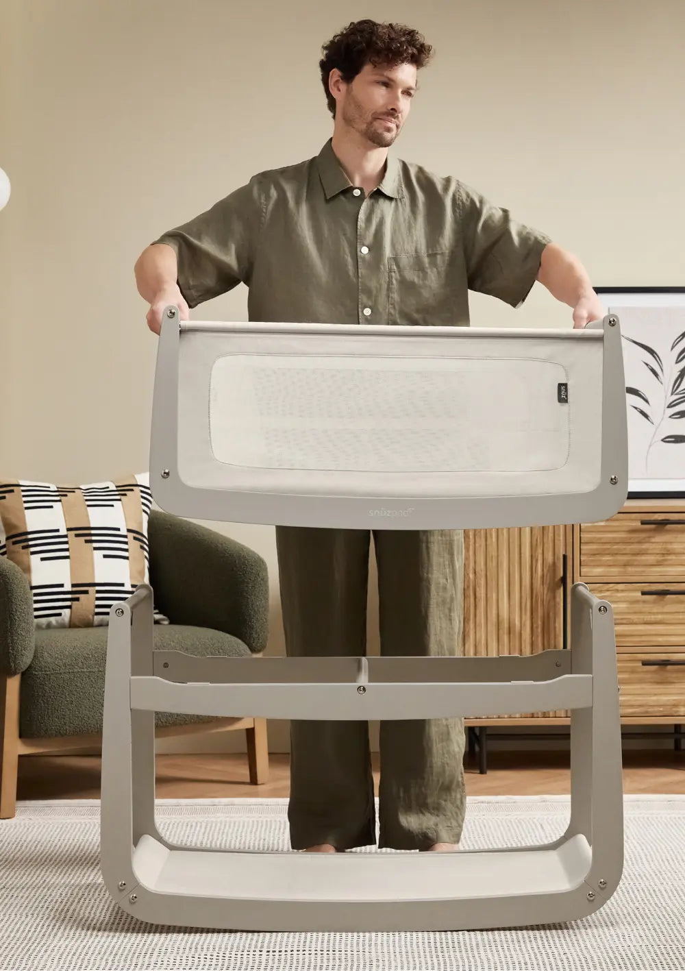 Ein Mann demonstriert die Funktion der abnehmbaren Liegewanne beim grauen Snüz Beistellbett SnüzPod 5 Pebble, indem er sie vom farblich passenden Holzgestell hebt.