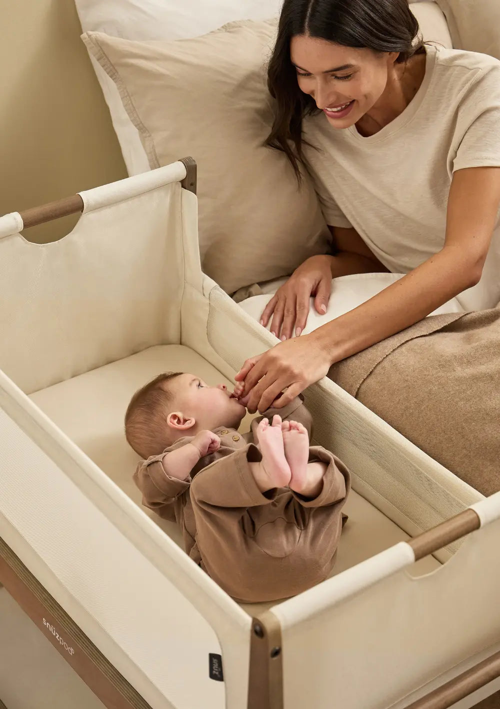 Eine Mutter liegt im Bett direkt neben dem geöffneten Snüz Beistellbett SnüzPod 5 Walnuss und hält zärtlich die Hand ihres auf dem Rücken liegenden Babys.