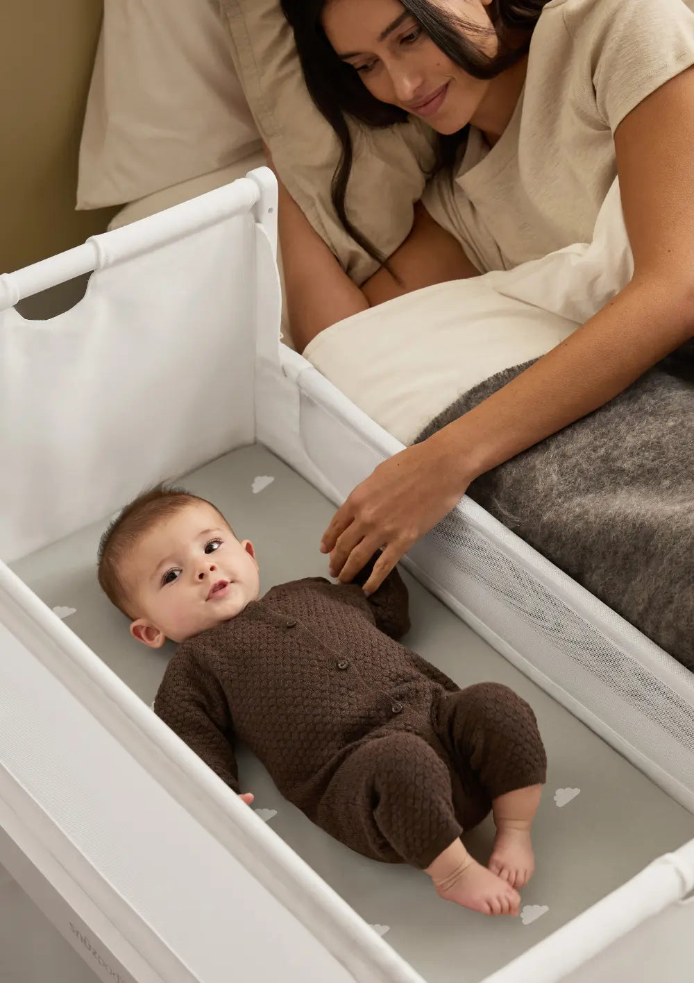 Sicheres Co-Sleeping: Eine Mutter liegt im Elternbett direkt neben dem geöffneten weißen Snüz Beistellbett SnüzPod 5 und hält die Hand ihres Babys.